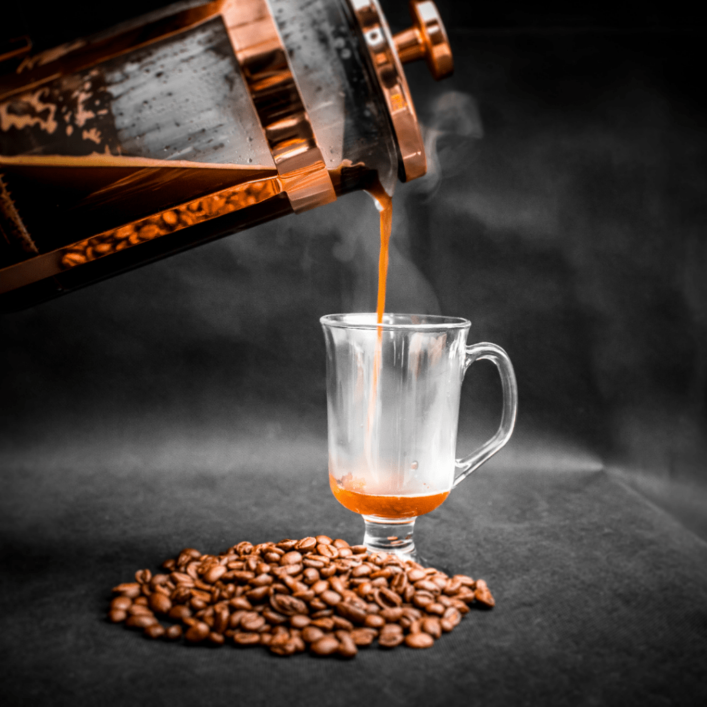 French press with bronze shell pouring into a glass with whole coffee beans in front of it